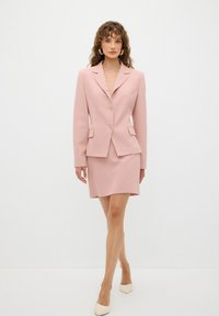 Blazer rosa claro con dos bolsillos frontales y falda corta a juego. Textura suave, corte entallado y cierre de botón sencillo. Tacones de punta.