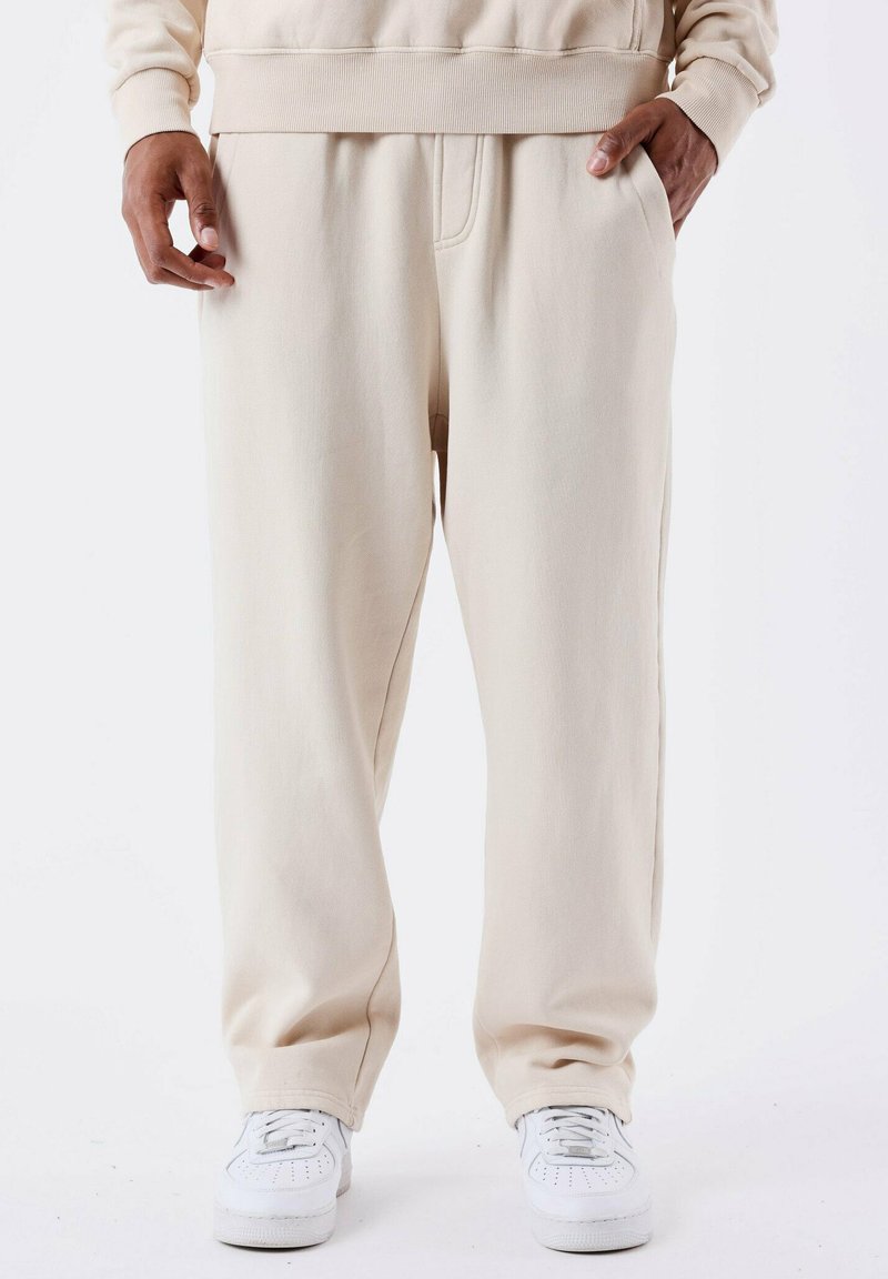 Beige joggingbroek van zacht materiaal, met een elastische tailleband en zakken aan de zijkant, gecombineerd met witte sneakers.