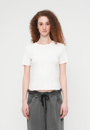 Top corto a maniche corte in maglia bianca a coste con bordi smerlati e una forma aderente, abbinato a pantaloni grigi con coulisse e tasche laterali.