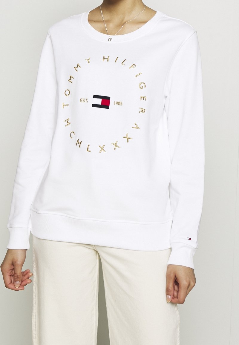 Personne portant un sweat-shirt blanc Tommy Hilfiger avec des lettres dorées et un logo circulaire, associé à un pantalon beige clair sur un fond uni.