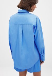 Light blue oversized shirt with long sleeves, buttoned cuffs, and a back pleat. Smooth texture, curved hem, and classic collar design.