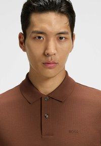 Brun polo skjorta med texturerad tyg, knäppkrage och broderad "BOSS"-logotyp. Har tre knappar och en slim fit-design.