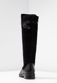 Bottes noires montant jusqu'aux genoux avec un tige en daim, des accents en cuir et une boucle décorative en haut. Bout arrondi et talon bas empilé.