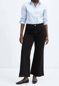 Persona vestida con una camisa de botones azul claro metida dentro de unos pantalones anchos negros y zapatos planos negros con tiras a los tobillos.