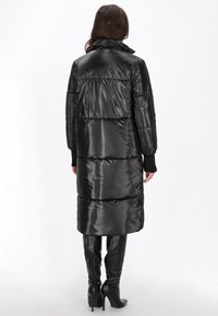 Sort pufferjakke med en blank, glat tekstur, der har en høj krave og en lang, løs pasform. Jakken har vandrette kviltede sektioner.