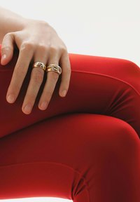 Dos anillos en una mano descansando sobre un tejido rojo; uno de oro con un diseño en eslabones, y otro de plata con un patrón texturizado. Piel suave visible.