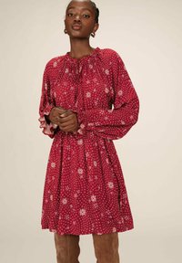 Robe rouge avec des motifs floraux, décolleté froncé, et manches longues avec des poignets à volants. Tissu lisse avec une coupe fluide et décontractée.