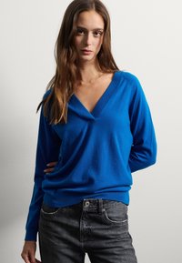 Jeune femme aux longs cheveux bruns portant un pull bleu à décolleté en V et un jean noir, se tenant contre un mur clair uni.