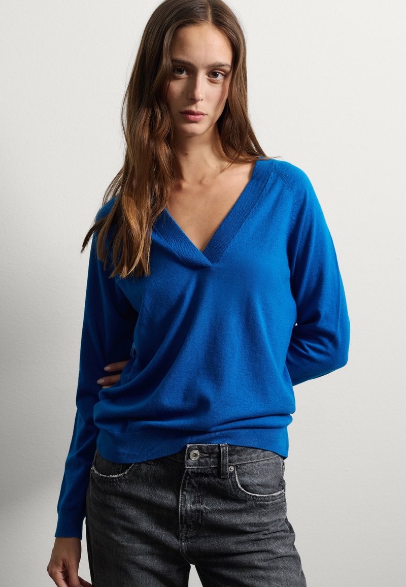 Jeune femme aux longs cheveux bruns portant un pull bleu à décolleté en V et un jean noir, se tenant contre un mur clair uni.