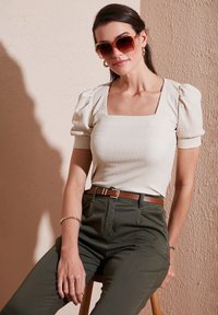 Haut beige à côtes avec manches bouffantes et décolleté carré, associé à un pantalon taille haute vert olive et une ceinture en cuir marron. Lunettes de soleil écaille.