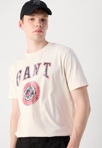 Cream cotton t-shirt featuring a red and blue "GANT" logo and circular graphic on the front. Styled with a black cap.