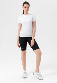 Hvit kortermet sport T-skjorte med logo, kombinert med svarte knelange sykkelshorts med en liten logo. Hvite sportsko.