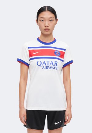 Jeune femme portant un maillot de football blanc du Paris Saint-Germain avec des bandes rouges et bleues et un short Nike noir, debout devant un fond uni.