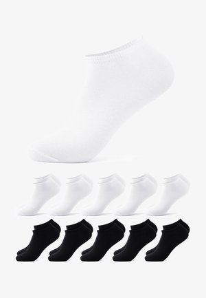 Person, die schwarze Knöchelsocken und schwarze Jeans trägt, steht auf einer weißen Oberfläche mit einem leicht erhobenen Fuß.