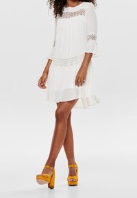 White dress with lace accents, pleated design, and bell sleeves. Worn with yellow platform sandals featuring ankle straps.