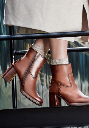 Bottines en cuir marron avec talons blocs et fermetures éclair latérales, portées avec des chaussettes beiges et un long manteau beige.