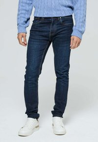Jean en denim bleu foncé avec une coupe slim, dotés de poches avant, de passants de ceinture et de détails de couture subtils. Associé à des baskets blanches.