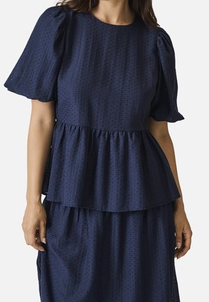Kobieta nosząca teksturowaną granatową sukienkę z krótkimi bufkami i marszczonym peplum w talii.