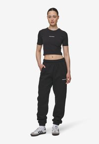 Černé cropped tričko s potištěným logem, v kombinaci s černými teplákovými kalhotami se stahovací gumou v pase a na nohavicích. Doplněno šedými sportovními botami.
