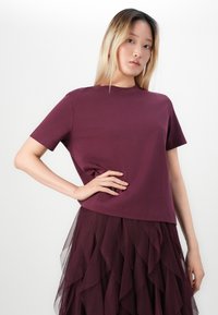 Giovane donna con capelli biondi lisci indossa una maglietta viola a maniche corte e una gonna a strati in rete viola, posando con una mano sui fianchi.