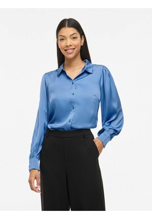 Femeie purtând o bluză strălucitoare albastră din satin cu mâneci lungi și pantaloni negri cu talie înaltă, zâmbind cu o mână în buzunar.