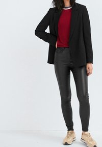 Blazer noir sur un t-shirt rouge à col rond, associé à un pantalon skinny noir et des baskets beige. Textures lisses et coupe ajustée sur l'ensemble.