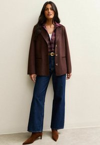 Blazer de cuero marrón con solapas con muescas, botones de un solo pecho, usado sobre una camisa de cuadros, combinado con jeans de cintura alta azules y tacones marrones.