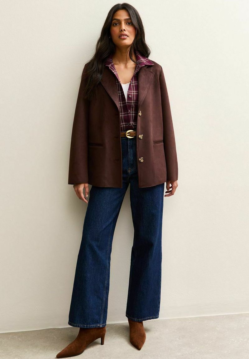 Blazer de cuero marrón con solapas con muescas, botones de un solo pecho, usado sobre una camisa de cuadros, combinado con jeans de cintura alta azules y tacones marrones.