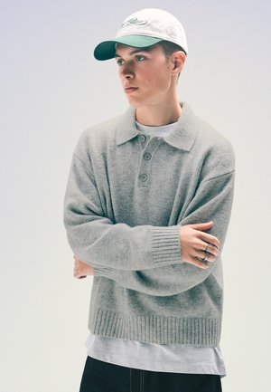Pull en tricot gris clair avec un col et trois boutons. Poignets et ourlet côtelés. Associé à une casquette blanche et verte.