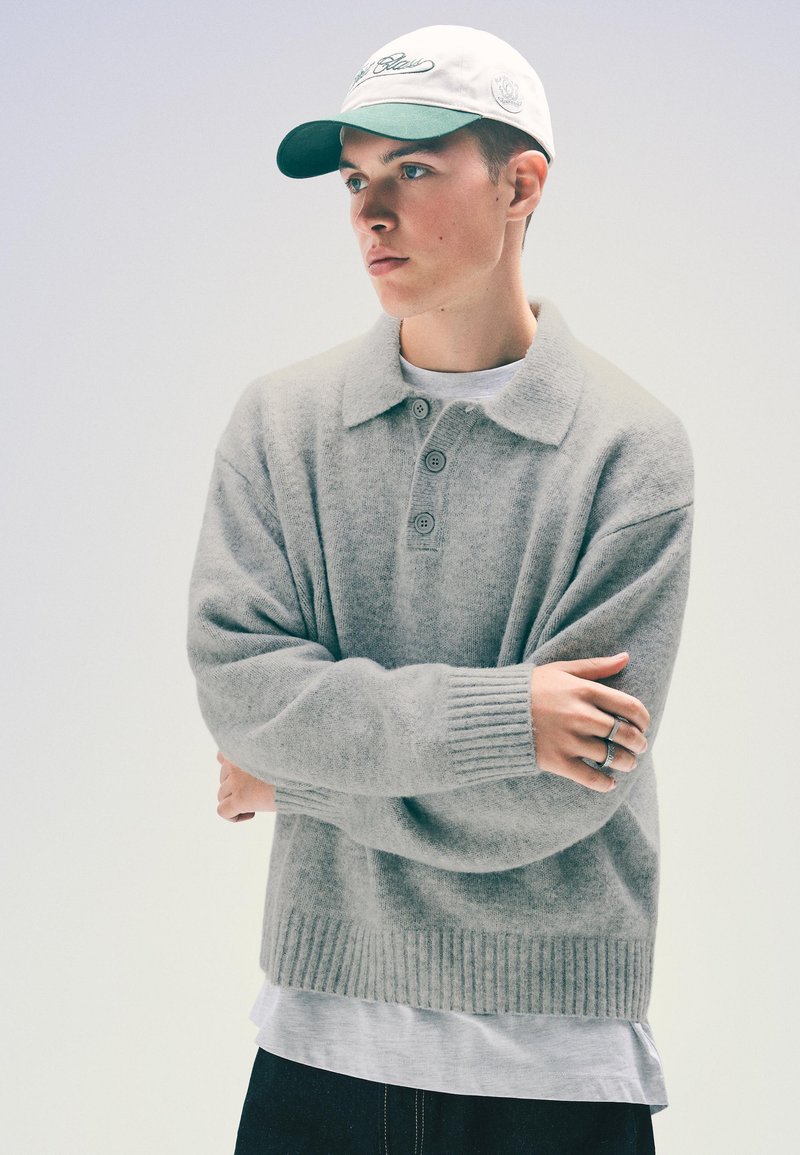 Világosszürke kötött pulóver, gallérral és három gombbal. Bordás mandzsetták és alj. Fehér és zöld sapkával párosítva.