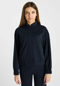 Marineblauwe ritsjack van zachte stof, met een staande kraag, elastische manchet en een subtiele logoprint op de borst.