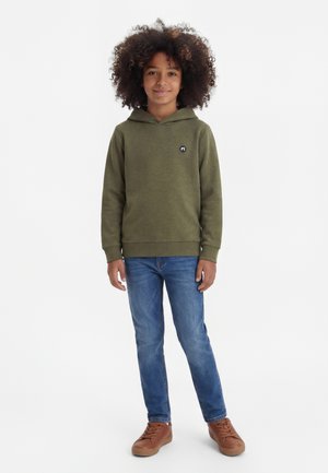 Enfant aux cheveux bouclés portant un sweat à capuche vert olive, un jean bleu et des baskets marron, debout devant un fond blanc.