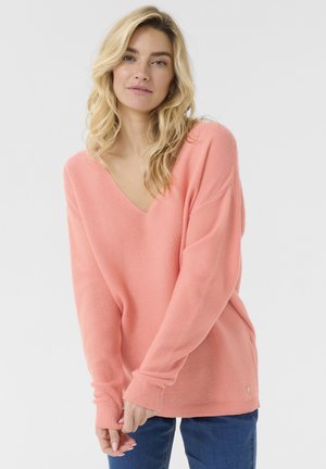 Lyserød V-hals sweater med ribbet tekstur, løs pasform og lange ærmer. Har en lille guldakcent ved falden. Bæres med blå jeans.