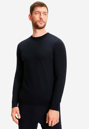 Mann mit kurzen braunen Haaren und Bart, der einen schlichten schwarzen langärmeligen Pullover und schwarze Hosen trägt und leicht zur Seite schaut.