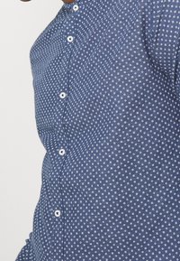 Sininen napitettu paita, jossa on pieni valkoinen geometrinen kuvio, päällä henkilö, keskittyen rinta- ja vartaloalueeseen.