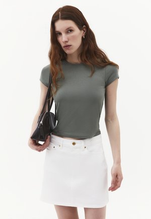 Giovane donna con lunghi capelli rossi che indossa una t-shirt grigia attillata e una gonna bianca, tenendo una borsa nera sulla spalla contro uno sfondo uniforme.