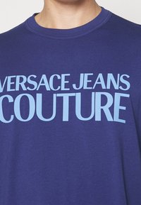 Camiseta de algodón morada con un estampado bold en azul claro que dice "VERSACE JEANS COUTURE". Diseño de cuello redondo con mangas cortas.