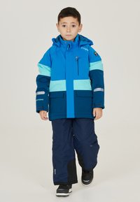 Blaue Kinderjacke mit hellblauen Streifen, einem Reißverschluss-Brusttasche und reflektierenden Akzenten, kombiniert mit marineblauen Schneehosen und schwarzen Schuhen.