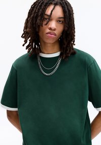 Mørkegrøn stof t-shirt med en løs pasform, der har en hvid rund hals og korte ærmer. To sølvkæder hænger omkring halsen.
