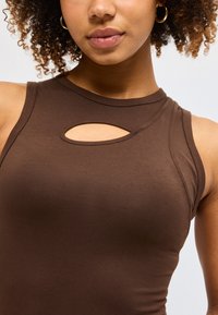 Νέα γυναίκα με σγουρά μαλλιά που φορά χρυσούς κρίκους στα αυτιά και ένα καφέ αμάνικο τοπ με μικρό οβάλ άνοιγμα κοντά στον λαιμό.