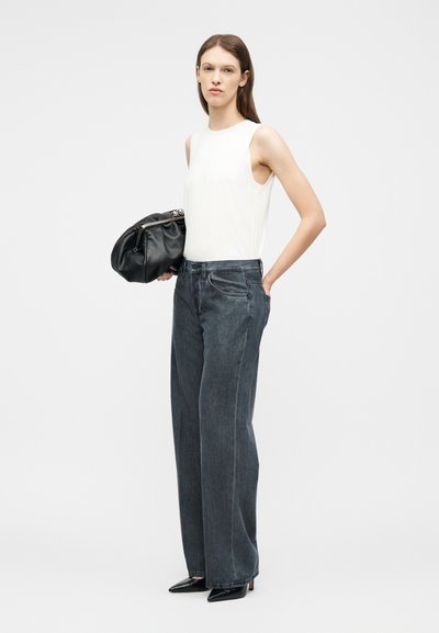 Femme portant un haut blanc sans manches, un jean large foncé, des talons noirs pointus, tenant un grand sac noir d'une main, l'autre main dans la poche.