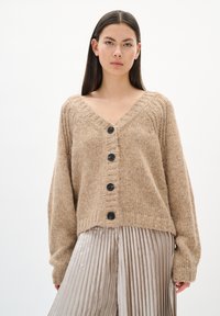 Beżowy sweter z dzianiny z głębokim dekoltem w serek, z pięcioma czarnymi guzikami i prążkowanymi detalami. Noszony na plisowanych metalicznych srebrnych spodniach.