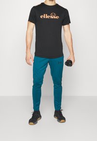 Man wearing black ellesse t-shirt, teal Under Armour joggers, black sneakers, holding black workout shaker bottle in right hand.