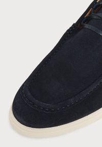 Gros plan sur une chaussure en suède bleu marine avec des coutures apparentes et une semelle en caoutchouc blanc, laissant apparaître une partie des lacets et de la tige.