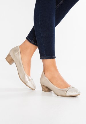 Ben iført mørkeblå skinny jeans og beige sko med lave hæle og strukturerede tåkapsler på et hvidt gulv.
