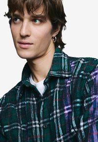 Grünes und schwarzes Flanellhemd im Karomuster mit weißen und lila Akzenten, ausgestattet mit durchgehender Knopfleiste und Kragen. Weiche Textur.