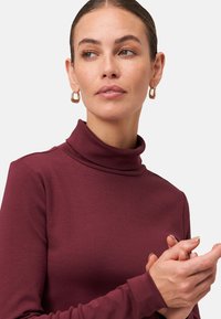 Woman with pulled-back hair wearing gold hoop earrings and a maroon turtleneck, looking to her left with hands clasped.