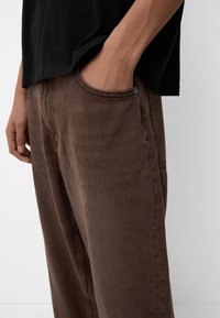 Brown denim jeans with a relaxed fit, featuring a classic pocket design and subtle stitching. Worn with a black shirt.