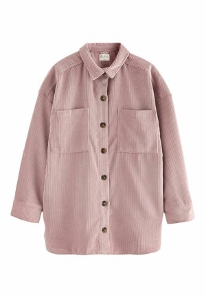 Femme portant une chemise en velours côtelé rose à boutons avec de grandes poches et un pantalon à imprimé léopard, debout à l'intérieur avec les mains sur les hanches.