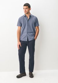 Chemise à manches courtes à motif bleu avec col boutonné, associée à un pantalon bleu marine et des chaussures marron foncé, portée par un mannequin masculin.